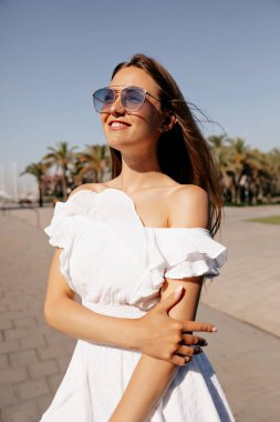 Beyaz yazlık elbiseli, güneş gözlüklü, açık havada poz veren neşeli gülüşlü romantik kadın.. 