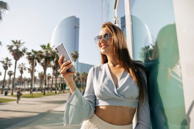 Image of cheerful cute woman holding phone and looking away on background of exotic sunny city . Smiling girl walking in the city with smartphone