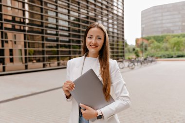 Dışarıda dizüstü bilgisayarı olan ve gülümseyen sevimli genç bayan. Güzel Avrupalı kız, günlük kıyafetler giyiyor, kameraya bakıyor ve çalışıyor.