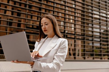 Beyaz ceketli, uzun saçlı, çekici bir kadın. Arka planda dururken dizüstü bilgisayar kullanıyor. Modern cam bina. . 