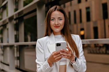 Avrupa 'da sokakta yürürken uzun düz saçlı, akıllı telefon kullanan Avrupalı çekici bir kadının açık hava portresi. Beyaz giysili güzel bayan dışarıda dinleniyor. 