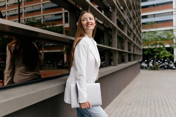 Smiling confident woman with long hair wraith white jacket is holding laptop and posing on background of modern business district in warm sunny day
