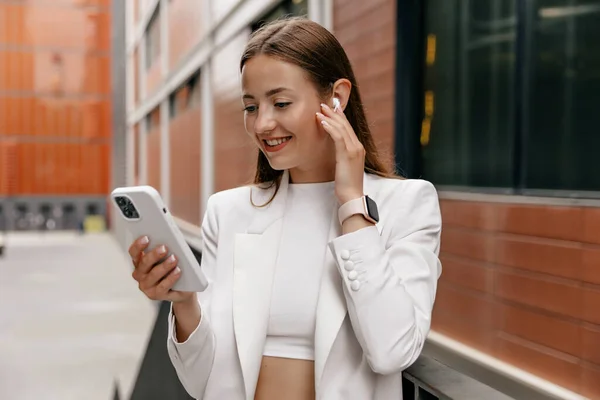 Stylish business woman with wonderful smile and light-brown hair wearing white jacket and shite shirt using smartphone and wireless headphones on business district