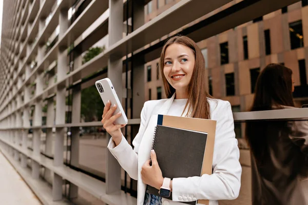 Attractive wonderful woman with long hair using smartphone and holding working notes on background of modern building in the city