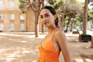 Attractive stylish european woman with dark hair wearing orange dress and head accessories posing outdoor on city park background 