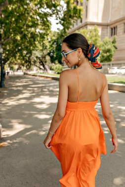 Close up portrait from back of adorable stylish slim woman with dark hair wearing orange dress and wearing head accessories walking on the street in summer park. High quality photo