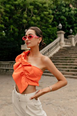 Good-looking european female model in pink fashion glasses wearing orange stylish blouse and beige jeans looking straight and walking outdoor in warm summer day.