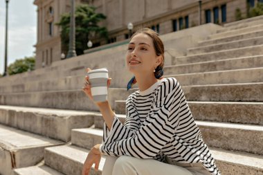 A beautiful young girl is enjoying a delicious coffee while sitting on the stairs. Attractive female model in a striped blouse is satisfied with a drink.