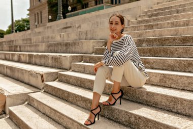 Tanned pretty brunette woman in beige pants and striped blouse poses sitting on the stairs. Young charming lady in stylish sandals with heels smiles.