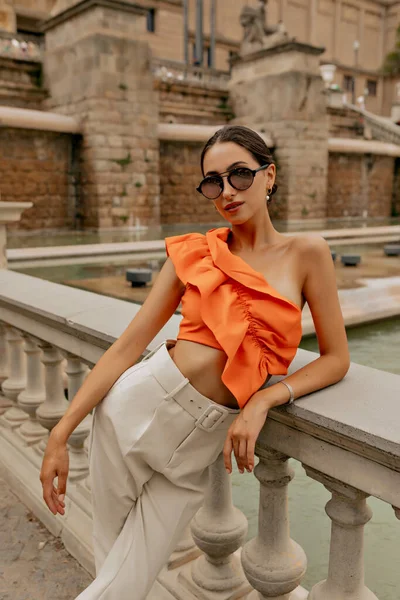 Charming young lady with dark hair is leaning on a parapet in stylish white trousers with a wide belt and orange blouse. Slender girl in stylish glasses against the background of historical place.