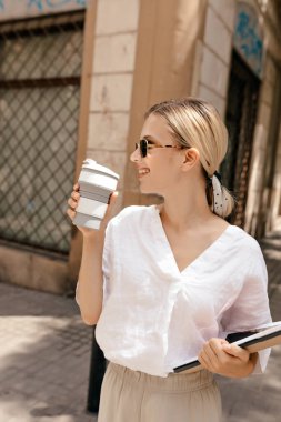 Lovely stylish young european woman with blond hair wearing white shirt and beige pants drinking coffee and walking around the street with notes. 