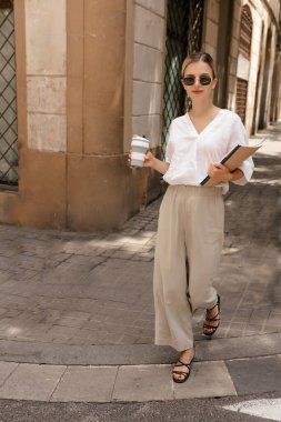 Full-lenght portrait of smiling european pretty girl in sunglasses walking with coffee and magazines in the city. Blond wearing white shirt and beige pants walking on heels