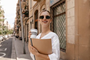 Happy and satisfied young girl with blond hair and smile enjoying a cup of coffee. Portrait of a beautiful blonde woman with notes and books in her hands.