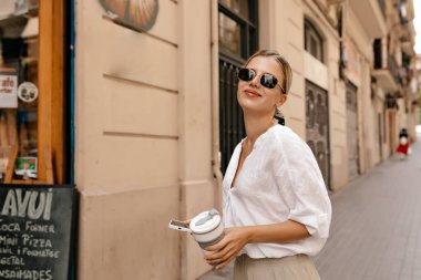 Satisfied young caucasian woman with snow-white smile looks at camera being outdoors. Stylish woman wears white shirt, sunglasses and ribbon on the hair. People emotions, lifestyle and fashion concept