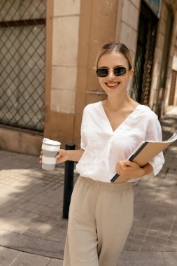 Smiling happy stylish woman in sunglasses wearing white blouse and beige pants in sunglasses walking on the street with coffee. Incredible stylish woman walking on work.