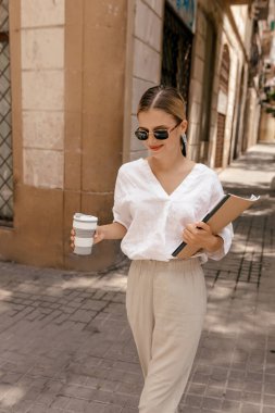Outdoor portrait of attractive stylish pretty girl with blond hair wearing white shirt and beige pants in sunglasses walking on the street in sunlight with notes and coffee. Fashion, beauty.