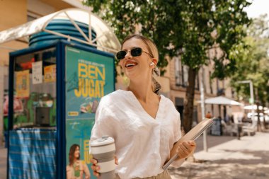 Smiling charming woman with blond hair in sunglasses wearing white shirt smiling and looking away with magazines and coffee in hands on city background in sunlight 