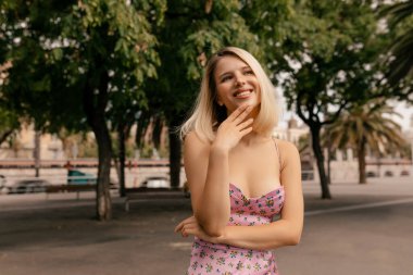 thoughtful romantic girl with short blond hairstyle in summer elegant dress touching her on chin and looking up with attractive smile outdoor on city park background in sunny day. 