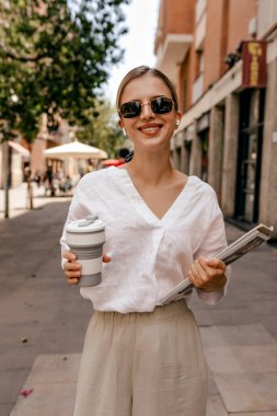 Attractive smiling caucasian woman with coffee and notes spends time outdoors. Happy girl wears casual clothes and sunglasses. Relaxed lifestyle, concept
