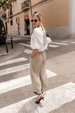 Happy charming successful woman with blond hair wearing white shirt and beige pants walking in the city in sunglasses and looking at camera with happy smile