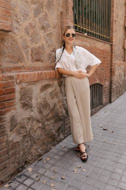 Full-length young girl in a good mood is walking around the city in black sunglasses. A cute blonde in a white blouse and beige pants with a ribbon in her hair.