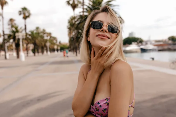 Cheerful young caucasian girl touching on chin and looking at camera spending leisure time outdoors during daytime. Blonde in sunglasses and dress walking on city with exotic plants on background