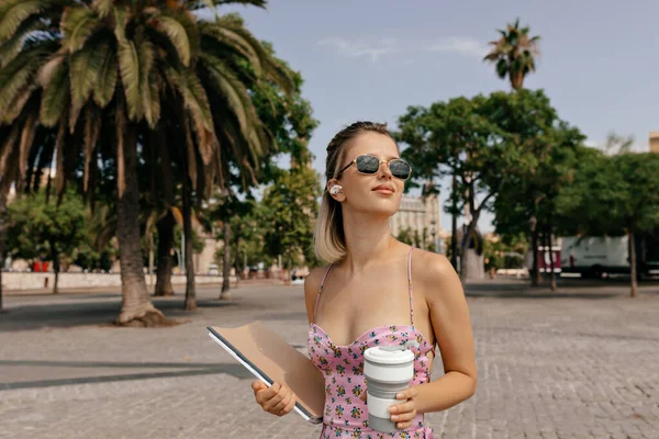 Elegant pretty woman with short hairstyle wearing pink summer silk dress walking on summer city with coffee and magazines on exotic plants background.