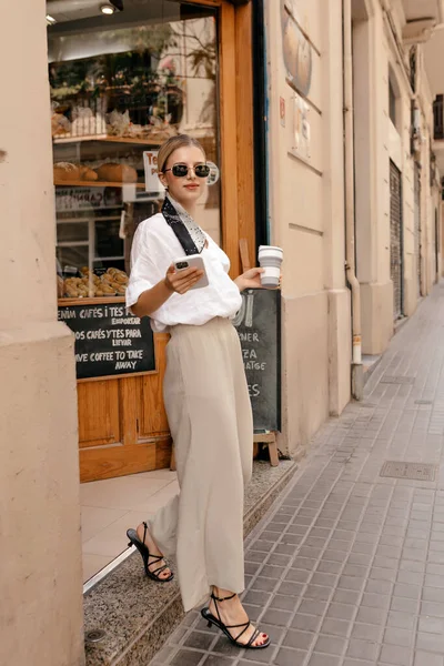 Full-lenght outdoor portrait of spectacular smiling woman wearing beige trousers, white shirt, sunglasses and ribbon on the hair is leaving bakery with coffee and holding smartphone in the hand. 