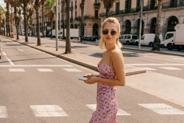 Elegant spectacular lovely woman with blond hair wearing pink summer silk dress and sunglasses walking on the street with exotic trees on background in sunny warm day.