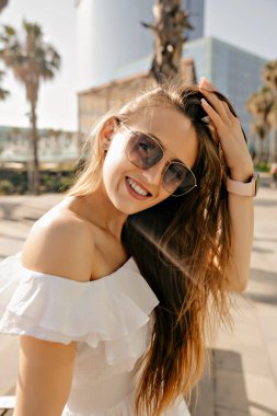 Stylish appealing woman with wonderful smile wearing sunglasses smiling at camera and touching her hair. Happy girl walking on quay with palms in sunny summer day
