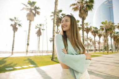 Cheerful caucasian girl laughing looking away spending leisure time outdoors in summer day. Long-haired girl keeps hands in her waist, wears shirt. Sincere emotions concept