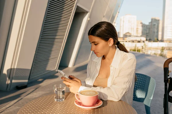 Focused attractive girl with dark hair and tanned skin sitting on summer terrace in sunlight and using smartphone. Outdoor portrait of stylish woman with coffee and phone in outdoor cafe.