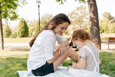 Pretty young woman with long dark hair sitting in the park on bright blanket with her little daughter and looking in smartphone. Woman spend time with daughter 