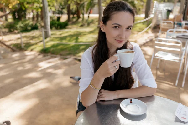 Adorable beautiful woman with dark hair sitting outdoor with coffee and looking at camera. Cute young caucasian brunette girl resting in park in summer day. 