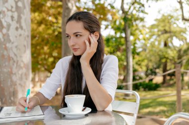Spectacular attractive woman with dark long hair wearing white shirt sitting outside in the park with cup of coffee and taking notes. Lifestyle concept