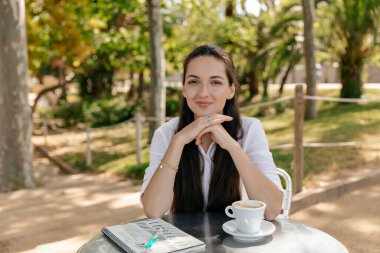 Smiling charming european woman with dark hair smiling at camera while sitting outdoor in park in sunlight. Lovely woman waiting for friends with coffee 