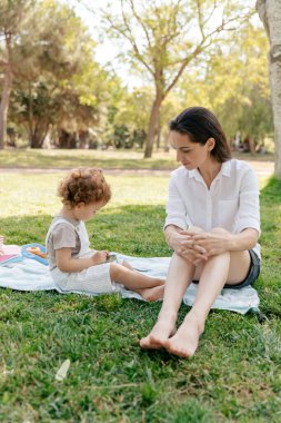 Full-length photo of young woman with dark hair wearing white shirt and shorts sitting in park with little daughter and having fun. 