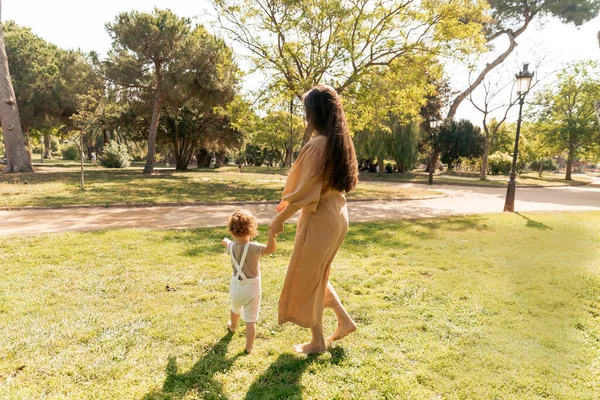 Full-lenght photo of long-haired woman in summer long dress walking in the park with her little daughter. Outdoor family photo of young woman with daughter 