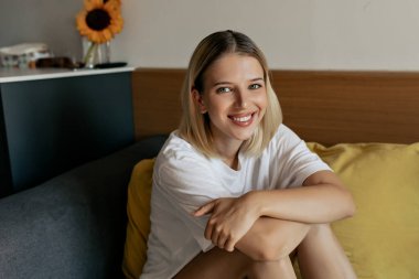 Smiling happy woman with blond hair, fairy skin posing at camera while sitting on the coach . Concept of working in home