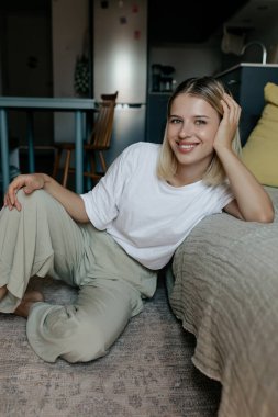 Smiling attractive woman with with short blond hair wearing home clothes sitting on the floor at home and posing at camera with wonderful smile. 