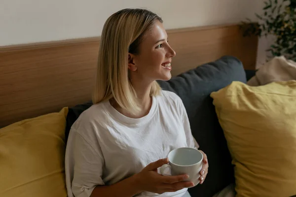 The side view of beautiful woman wake up at home . Caucasian young woman sitting on the coach with coffee , wearing white shirt. Concept of working in home