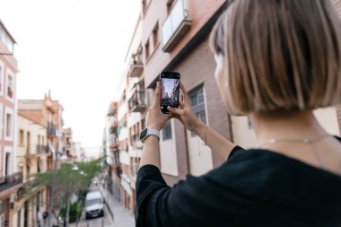 Kısa saçlı bir kadının arka tarafındaki portre günbatımında şehrin fotoğrafını çekiyor. Kafkasyalı sakin bir kız akıllı telefon ekranına bakar ve şehirde vakit geçirir. teknoloji kavramı