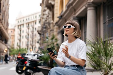 Avrupa görünümlü genç kadın, kentte eski Avrupa caddesinin arka planında kahveyle oturuyor ve akıllı telefon kullanıyor. Beyaz tişört, kot pantolon ve parlak güneş gözlüğü giyiyor.