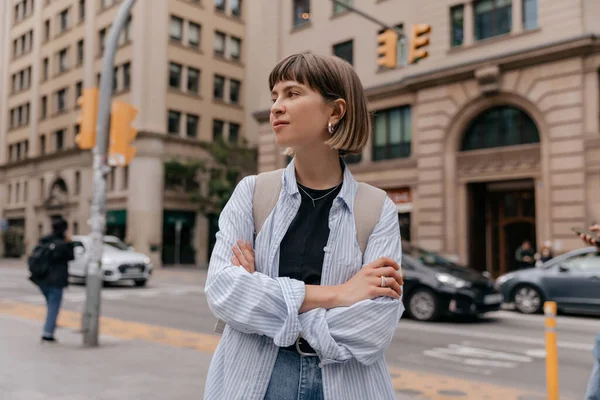 Outdoor photo of stylish european woman with short hair wearing blue shirt walking in the city with backpack. Attractive good-looking girl spend time in European city