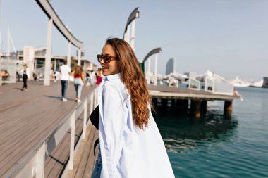 Güneşli bir günde sırtüstü iskelede yürürken gülümseyen mutlu kadının fotoğrafı. Esmer gülümser, günlük kıyafetler giyer. Samimi duygular yaşam tarzı konsepti. 