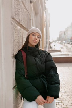 Şen şakrak bir kız, gün ışığında duvarın yanında poz veriyor. Bol kotlu, siyah ceketli ve gülümseyen bej şapkalı kadın.