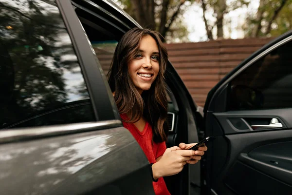 Attractive woman in red knitted shirt sitting in car with happy smile and using smartphone in the city. Brunette girl sits in grey car.
