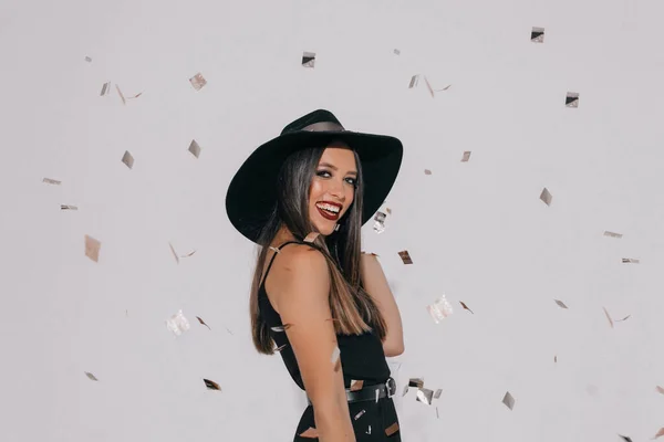 Stylish european woman with black hair wearing black hat and dress posing with adorable smile to camera over isolated background under confetti. Holiday, halloween, birthday.