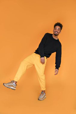 Uzun, yakışıklı, neşeli afro adam kameraya mutlu bir gülümsemeyle bakıyor. Koyu renk saç stili ve gösterişli kıyafetler stüdyoda ışık altında poz veriyor..