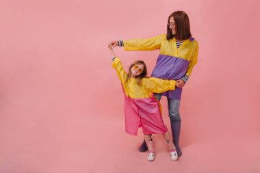 Çok güzel gülümseyen ve küçük kızıyla oynayan bir portre. Yağmurluk ve bot giyiyorlar. Küçük kızı olan genç bir kadın stüdyoda pembe arka planda poz veriyor. 